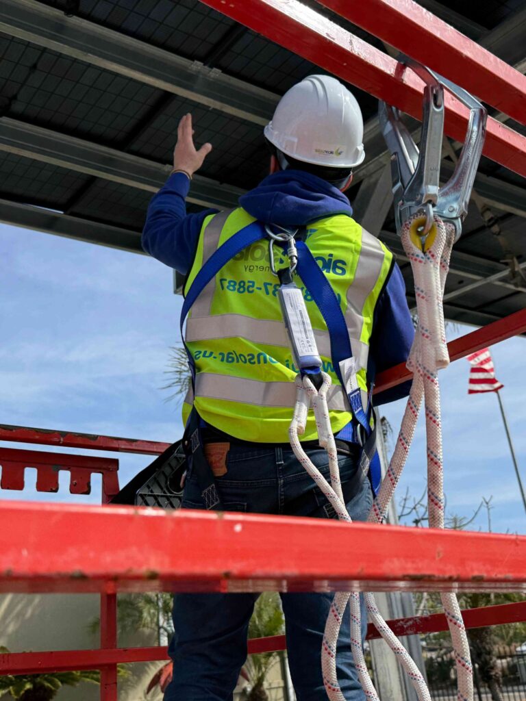 Solar technician on red scaffolding wearing safety gear and helmet.